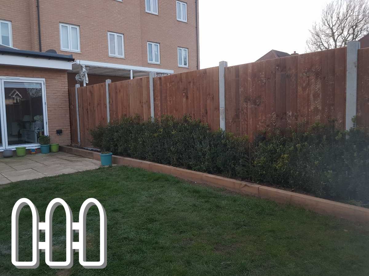 Residential garden with a neatly installed wooden fence featuring vertical planks and white concrete posts, complemented by lush green shrubs in a raised bed, and a tidy patio area.