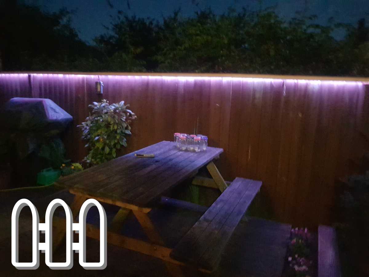 Nighttime view of a garden featuring a wooden fence illuminated by purple LED lights with a wooden picnic table and lush greenery.